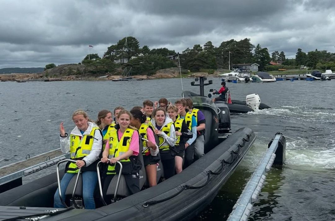 Smilende ungdommer i gule redningsvester sitter i en RIB-båt ved brygga, klare for tur