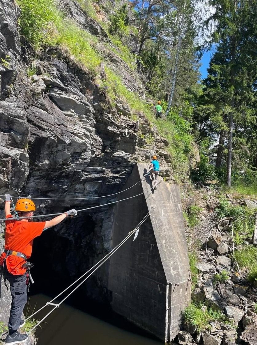 Ungdom i orange t-skjorte og klatrehjelm balanserer på en vaier over vannet ved fjellveggen på via ferrata, mens to andre klatrer videre opp